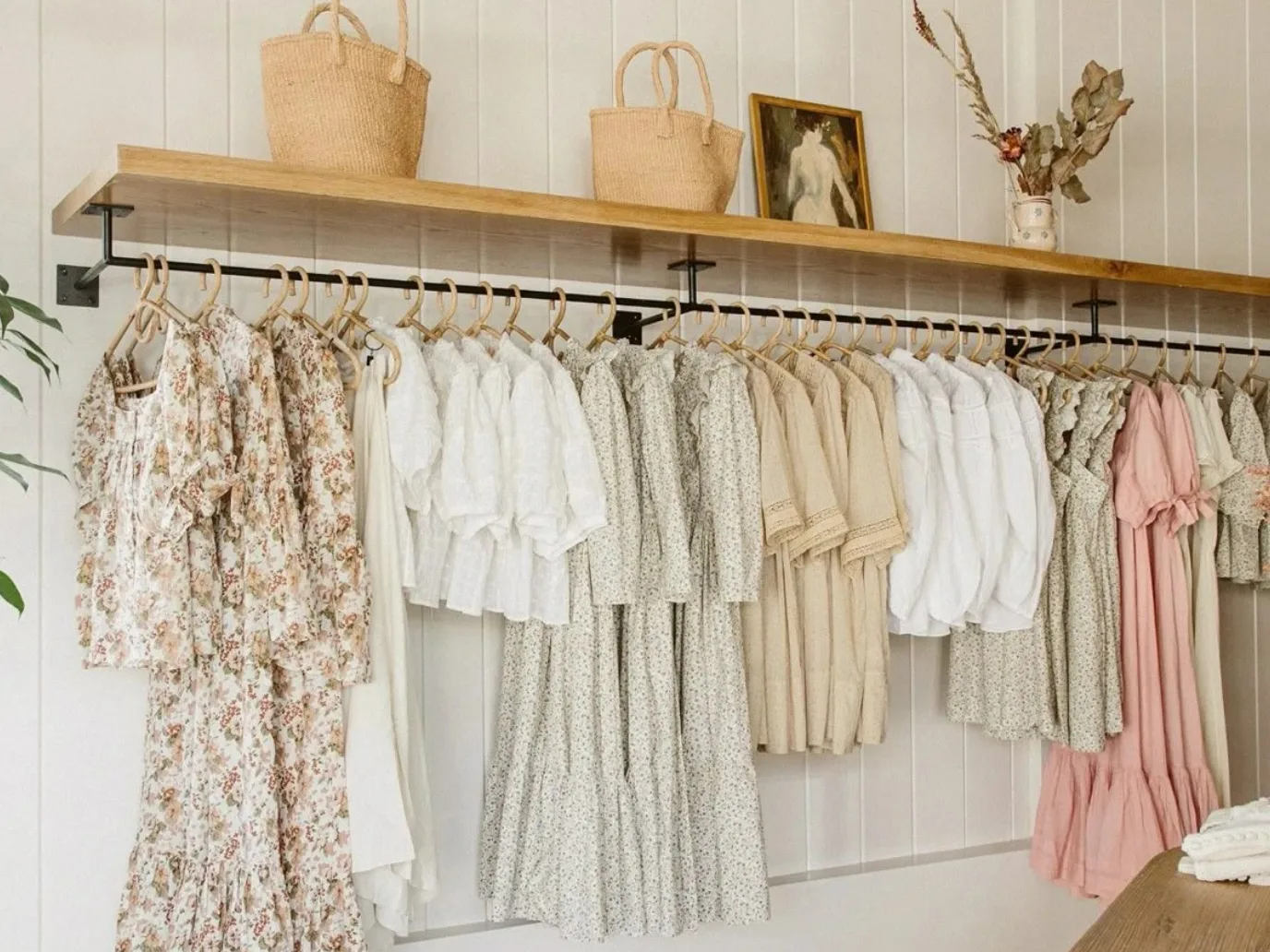 A boutique rail hung with floral and linen dresses in cream, sage, and blush, under a shelf of woven baskets