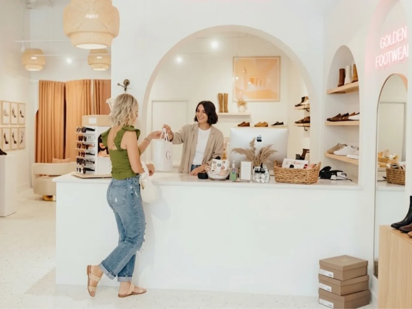 A shoe store associate handing a shopping bag across the counter to a customer, with boots and sneakers on arched shelves behind the register
